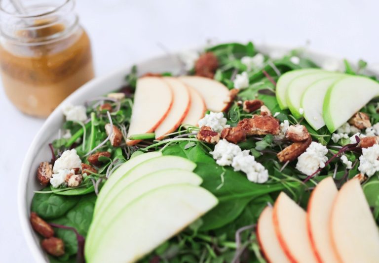 Bowl of baby spinach leaves with sliced apples, pecans and blue cheese crumbles with a small jar of vinaigrette next to it