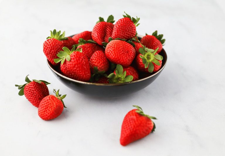 A bowl of whole strawberries sits in a black bowl with a gold rim. There are two strawberries to the bottom left of the bowl, and one strawberry slightly out of focus to the bottom right of the bowl. The background is greyish white marble.