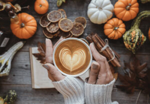 two hands holding a latte with a heart-shaped foam surrounded by small pumpkins and cinnamon