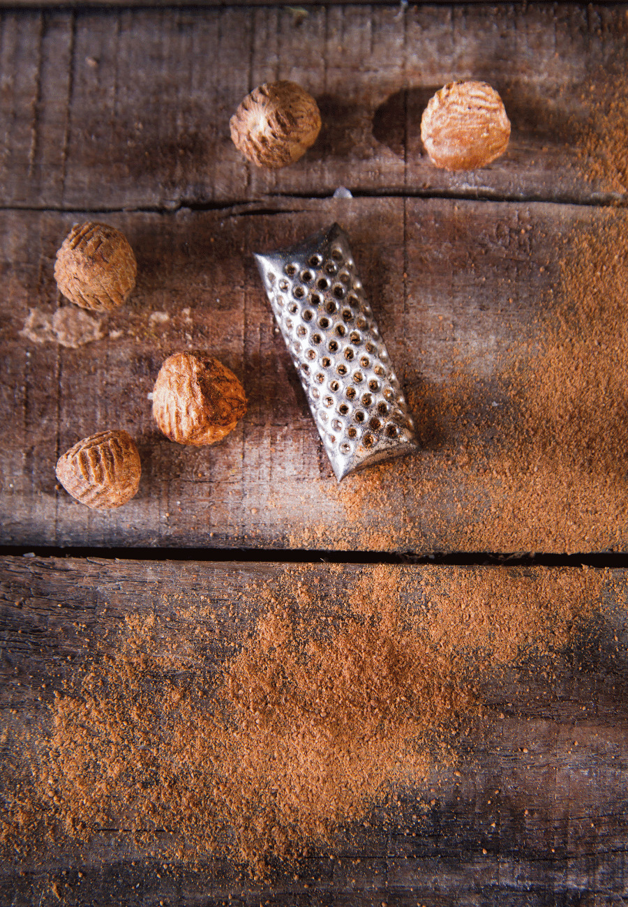 A handheld spice grater with whole nutmeg pods and ground fresh nutmeg on a wooden table