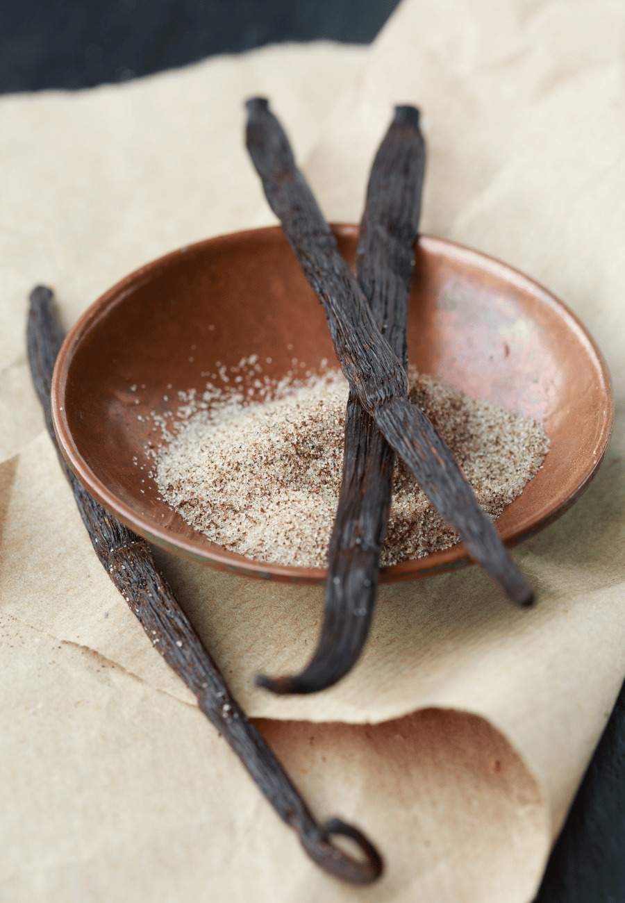 three vanilla bean pods resting on and near a small wooden bowl with a linen tablecloth