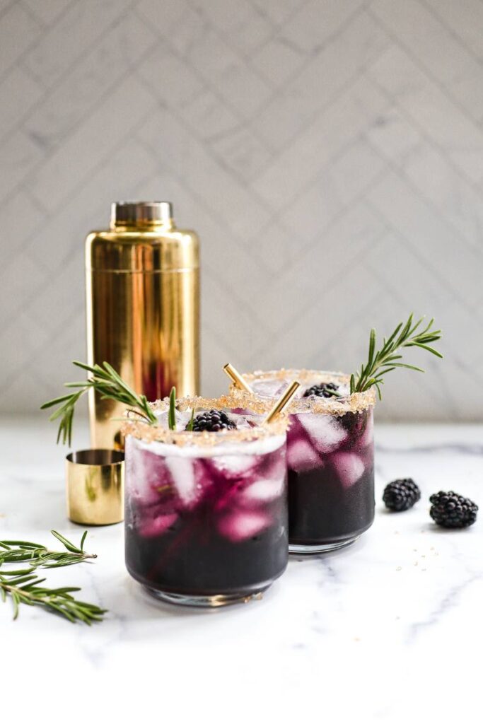 Two glasses with a deep purple mocktail and a gold cocktail shaker against a white background