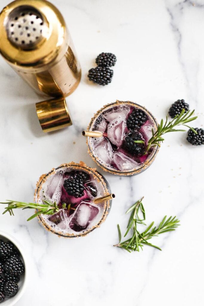 Overhead view of a purple mocktail filled with ice, topped with a fresh blackberry and rosemary sprig.