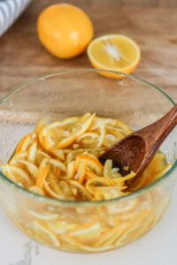 Sliced lemons being tossed in a clear glass bowl with honey and sugar, looking glossy and syrupy.