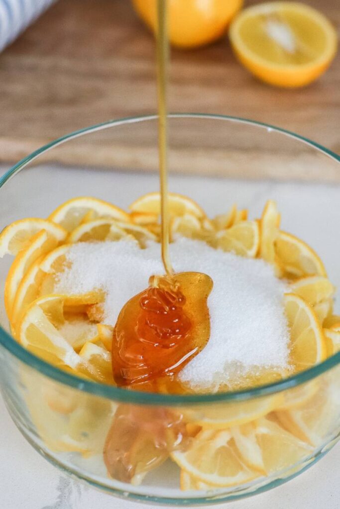 Pouring honey into a clear glass bowl that holds the thinly sliced Meyer lemons and sugar.