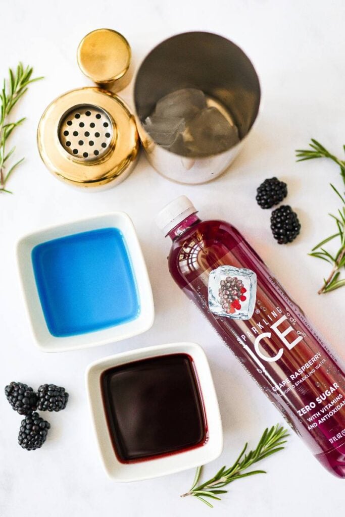 Ingredients for a purple mocktail laid out on a white surface: Blue Curacao syrup, grape juice, Sparkling Ice, and fresh blackberries.