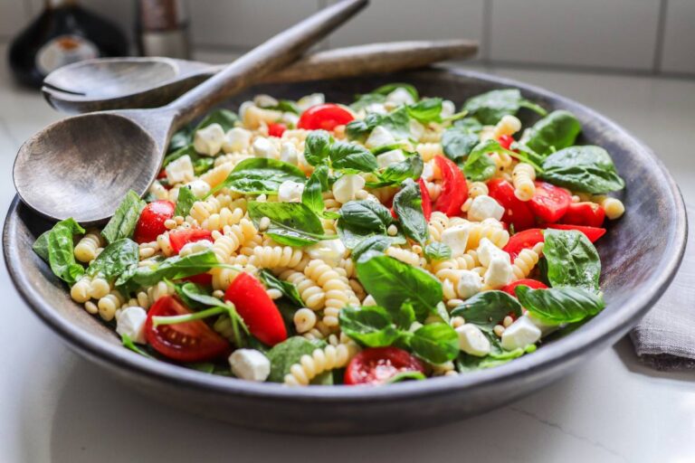 Large brown wooden bowl filled with pasta, green basil and red tomatoes with a serving set