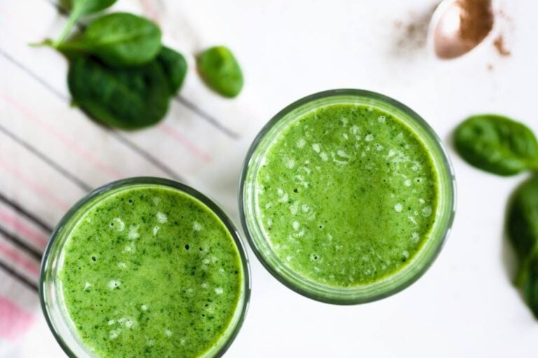 Overhead photo of two tall glasses of vibrant green detox juice with a fresh spinach on a bright white counter.