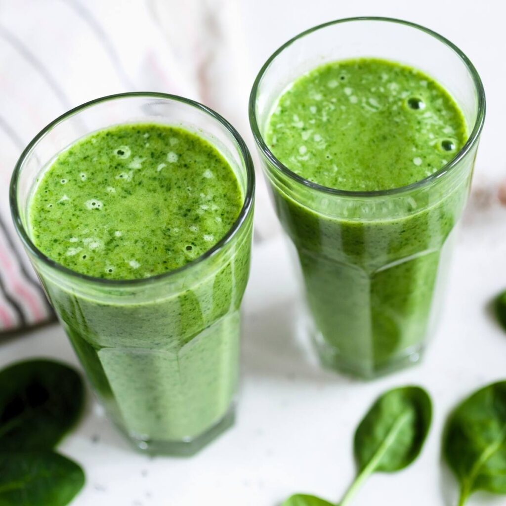 View of two tall glasses of green juice surrounded by fresh spinach with a striped tea towel in the background.