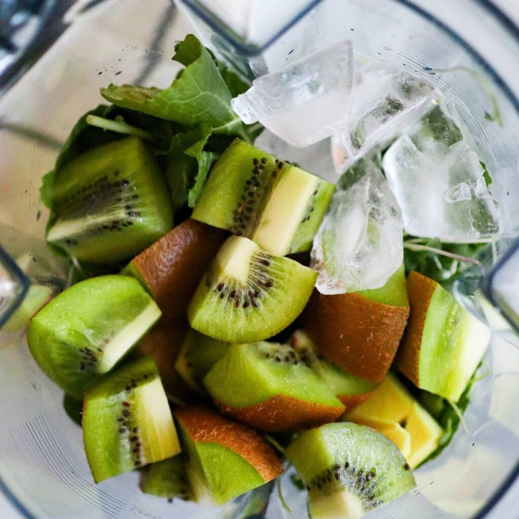 Adding ice and fresh kiwi to the greens in the base of a blender.