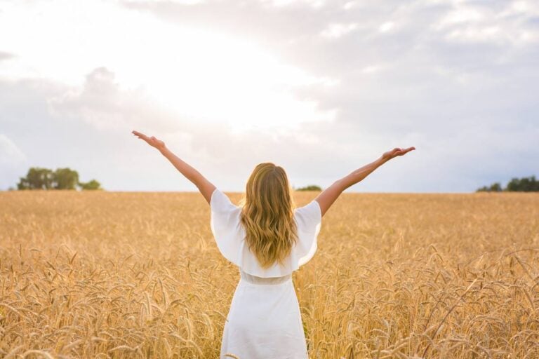 A close-up of a woman in a white dress standing in a field of golden wheat stalks in a sunlit field, illustrating the biblical theme of harvest and provision.